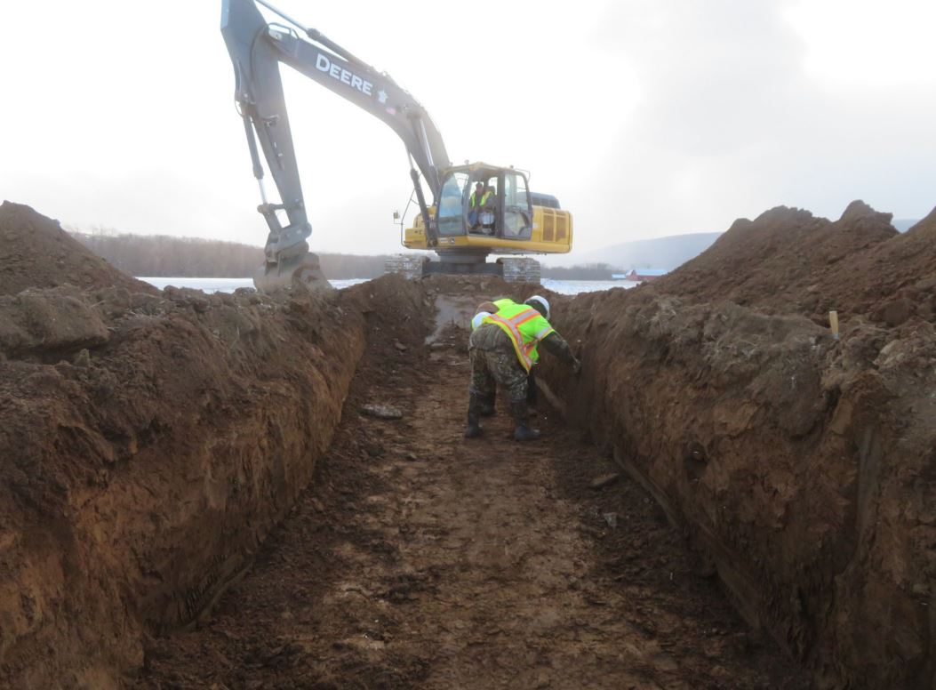 Man stands in large trench being excavated by machine.