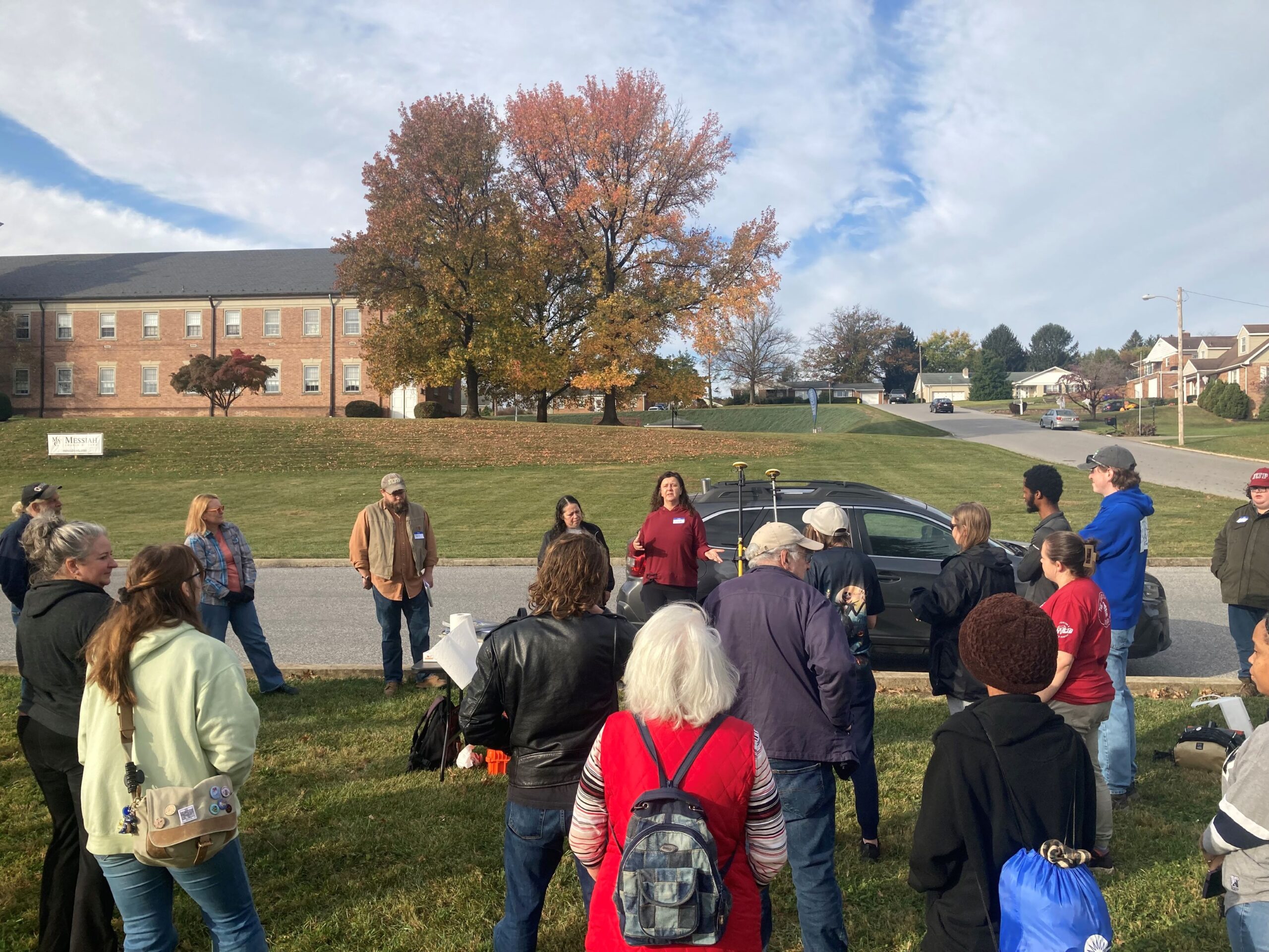 Two people address crowd of people standing on grass next to a road.