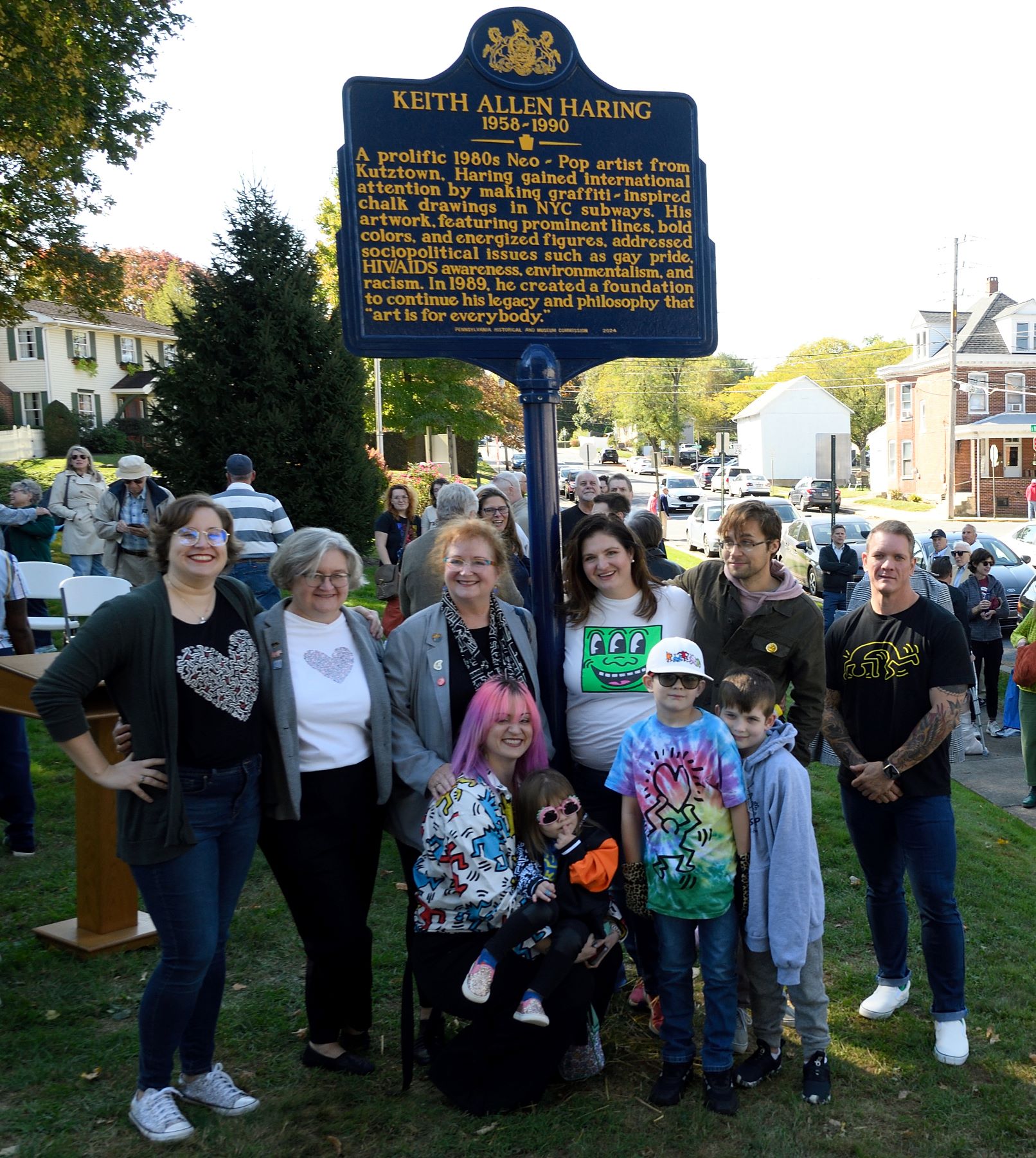 Group of people standing around tall metal sign on crowded lawn.