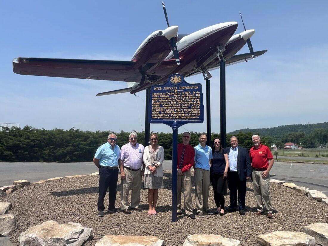 Group of people stand around a tall metal sign below a large model airplane.