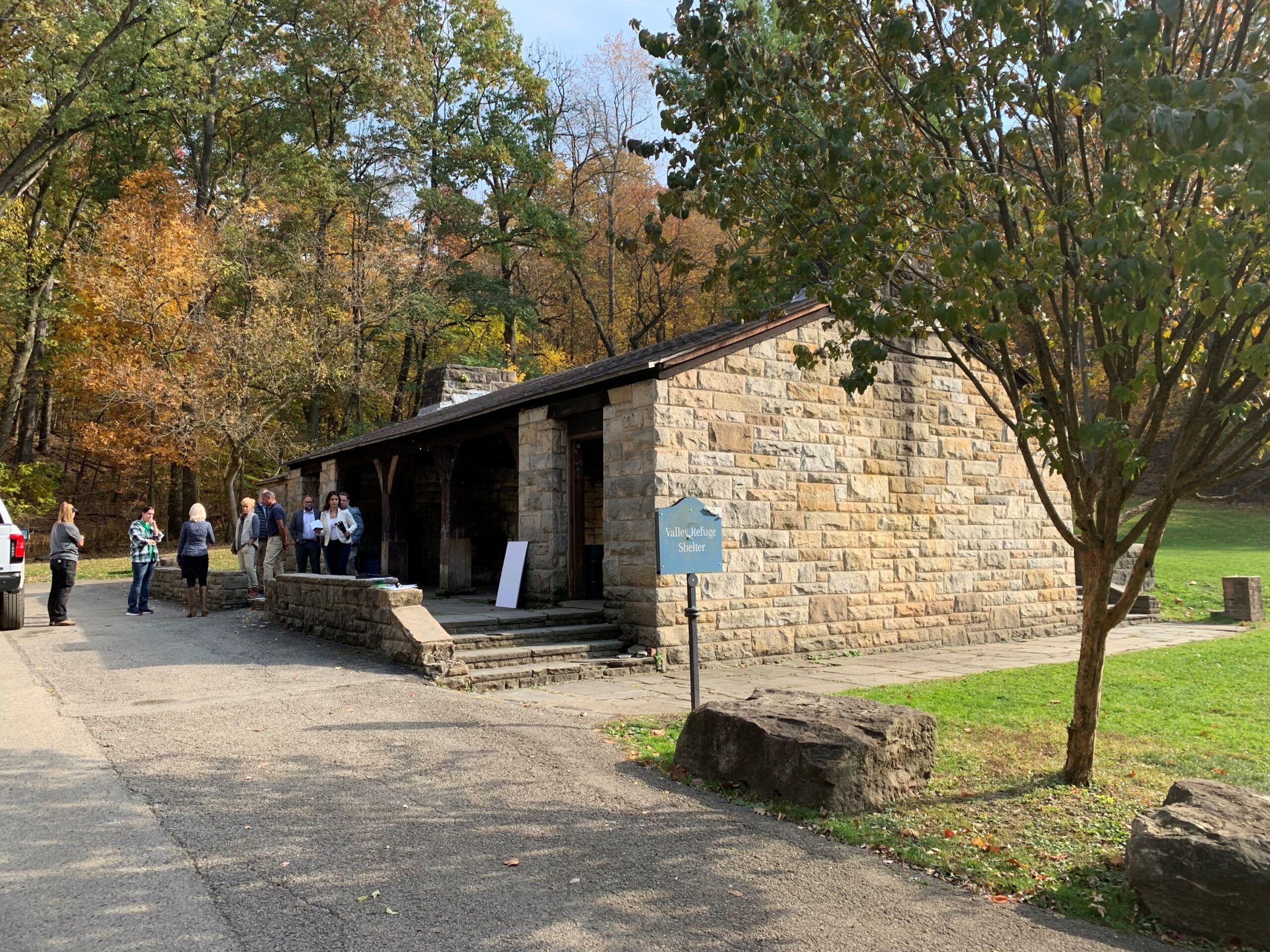 One-story stone building along a paved road and surrounded by grass. People are gathered at one end.