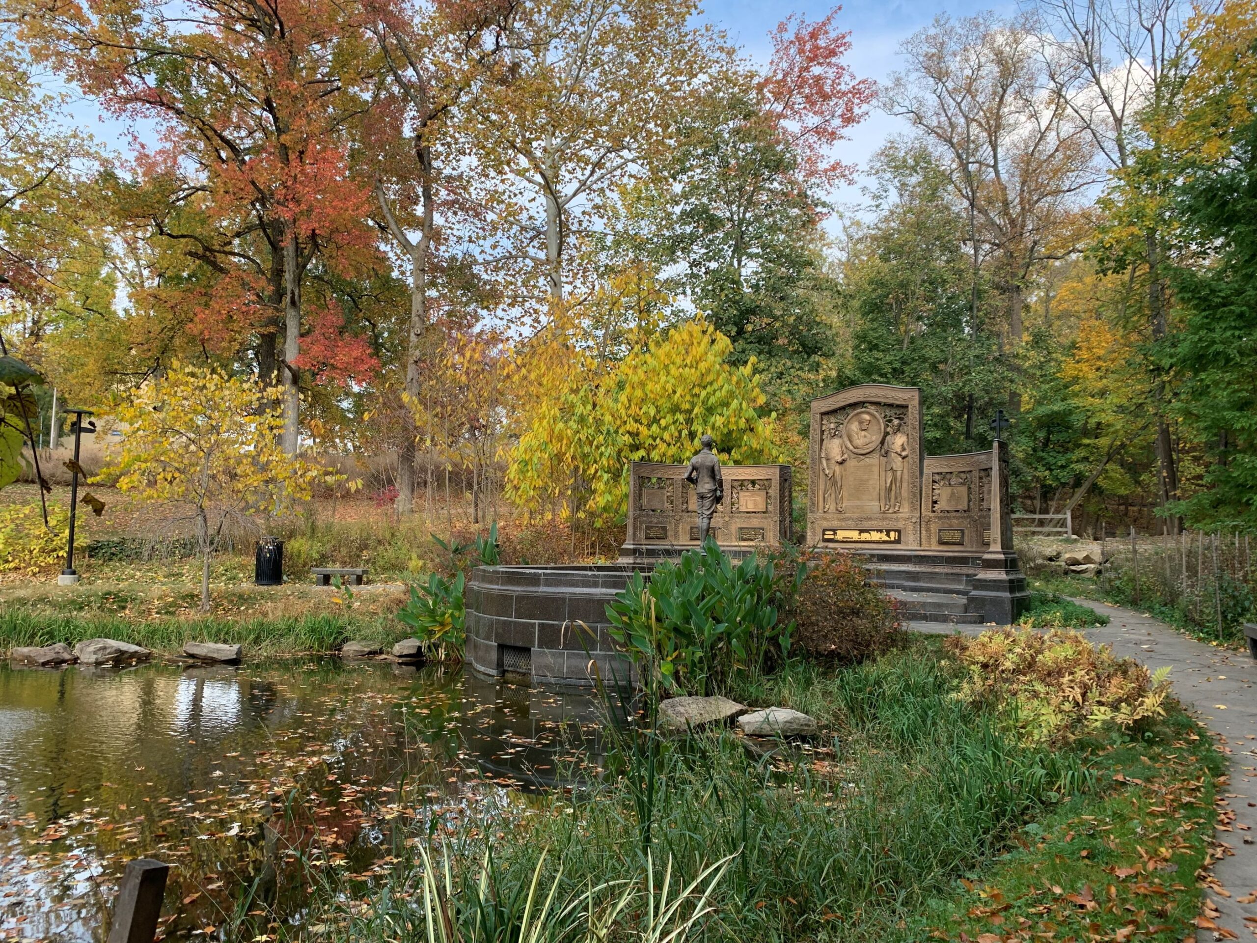 Group of masonry panels, steps, and statute between a paved path and water.