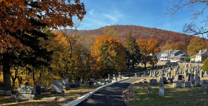 Looking across a cemetery with a paved road and many masonry grave markers on a hillside surrounded by wooded areas, homes, and mountains.