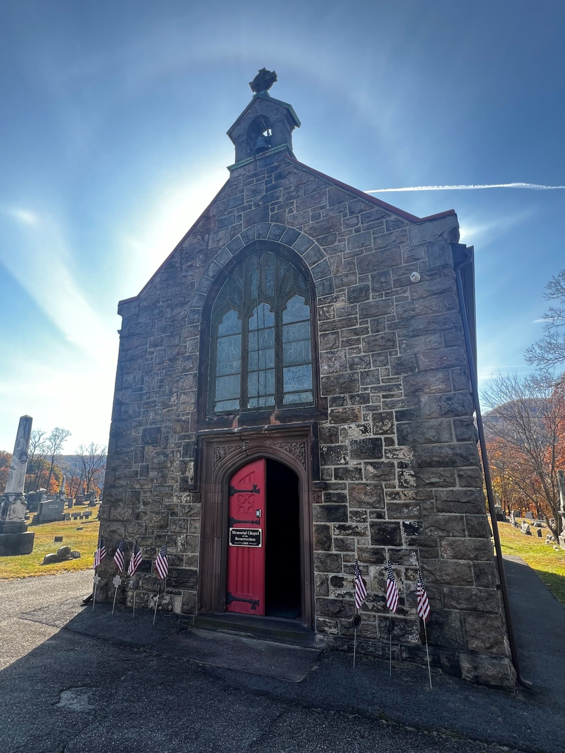 Small stone building with red doors, intricate details, and a large stained glass window surrounded by grass and stone grave markers.