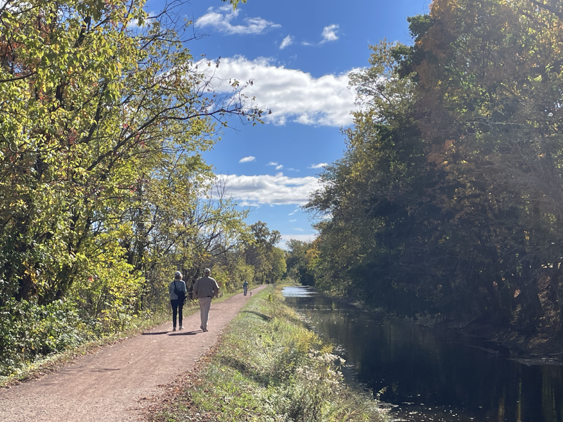 Two people walk along a dirt path next to a narrow body of water.