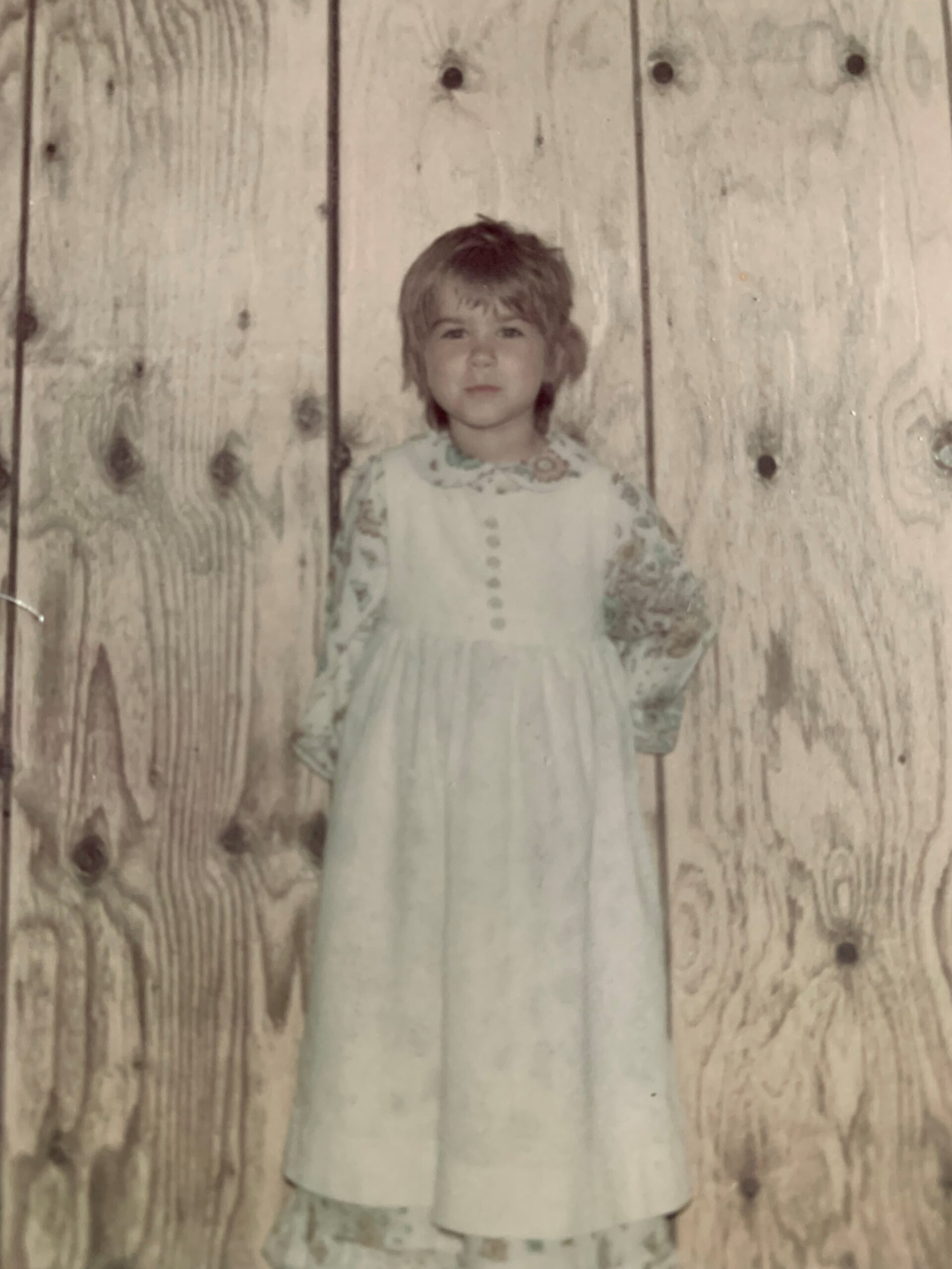 Small child in a dress standing in front of a wall.