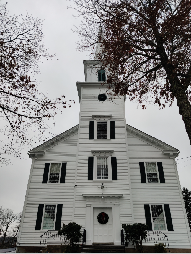 Two story church with central bell tower covered in wood clapboards with many multi-lite windows on both floors.