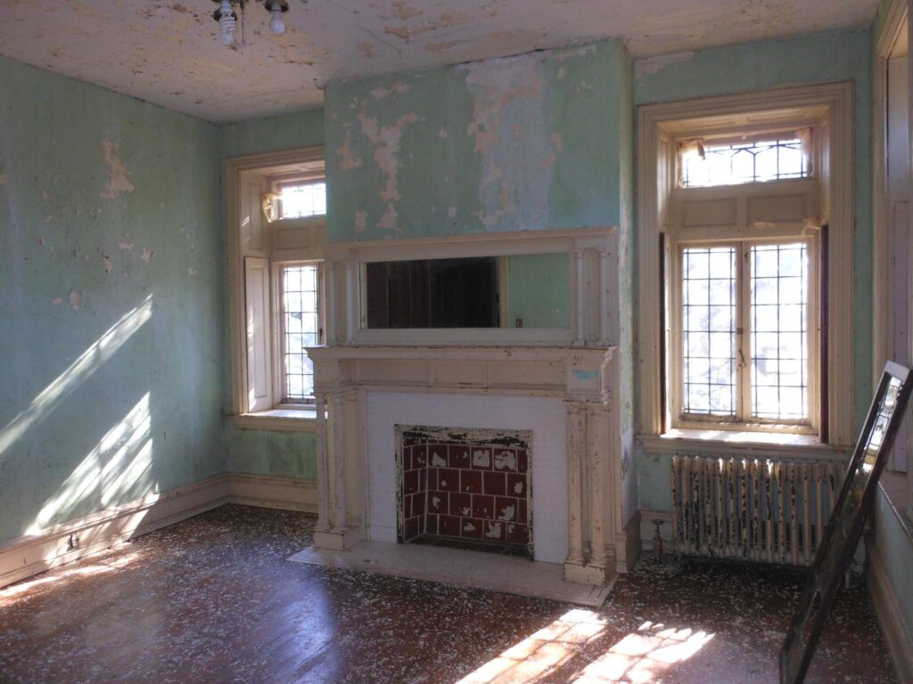 Room with flaking plaster and a fireplace with mirror on an exterior wall between two windows.