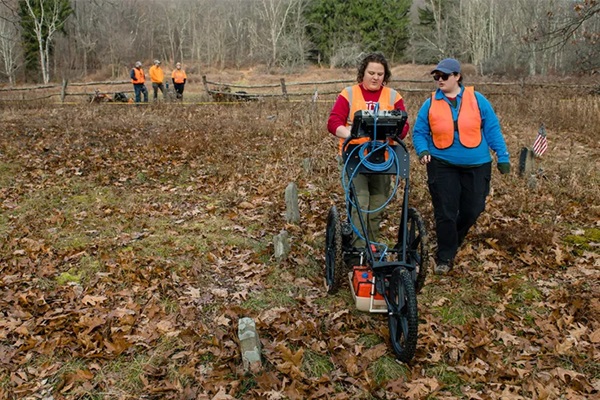 One person pushes a machine while another walks beside in a field in a wood area covered with leaves.