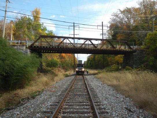 Railroad tracks run in a straight line below a metal truss bridge. A truck is parked on the tracks.
