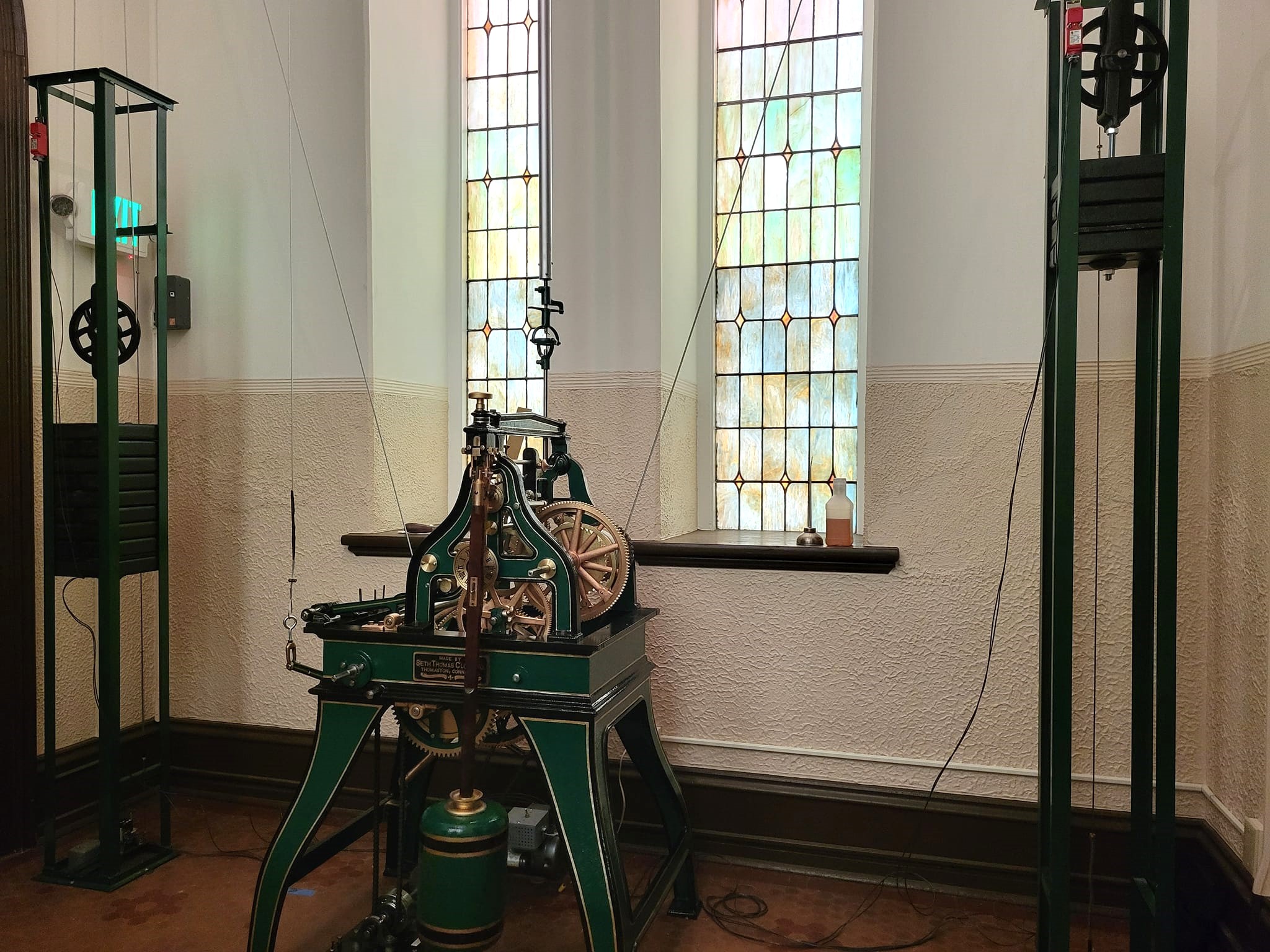 Clock mechanism inside a room in front of stained glass windows with wire connecting it to weights