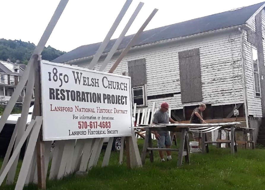 Two people standing outside a small wood building working on siding, with piles stacked around them.