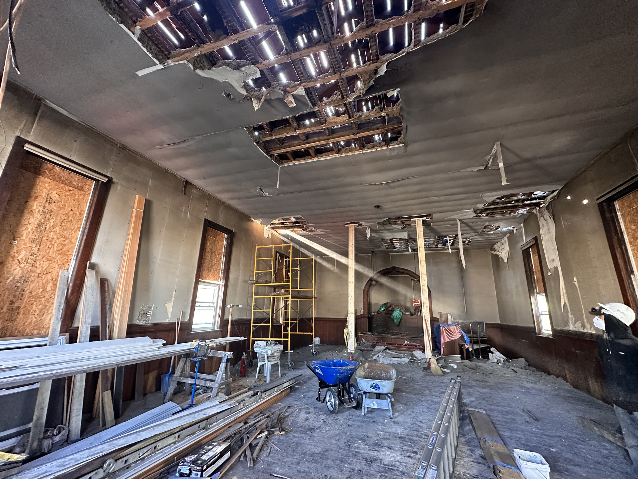 Large open room with dusty floor, boarded windows, plaster walls, holes in ceiling, and construction materials.