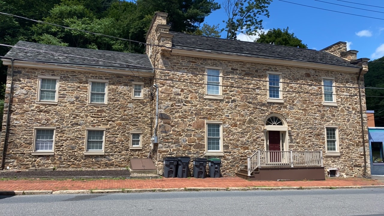 Large two-story stone house with stepped gable ends, center entrance, and many windows at the edge of a paved street.