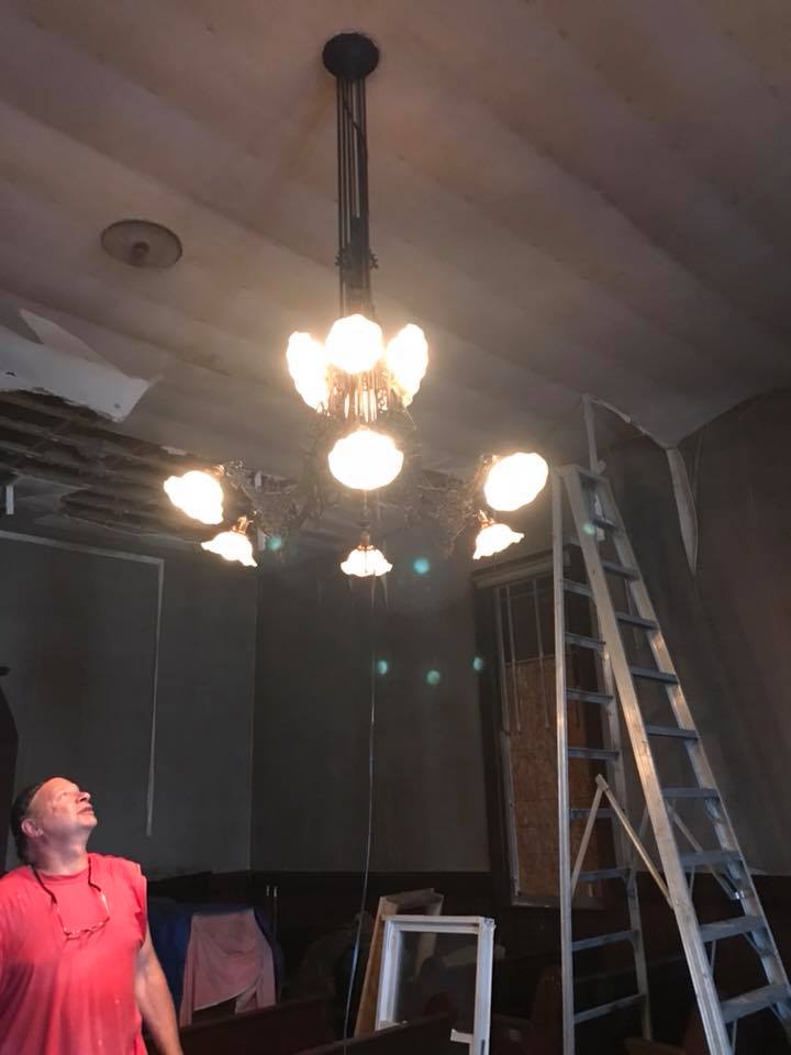 Man in red looks up at a lit chandelier in a dim room, with an aluminum ladder nearby. The ceiling shows signs of renovation.