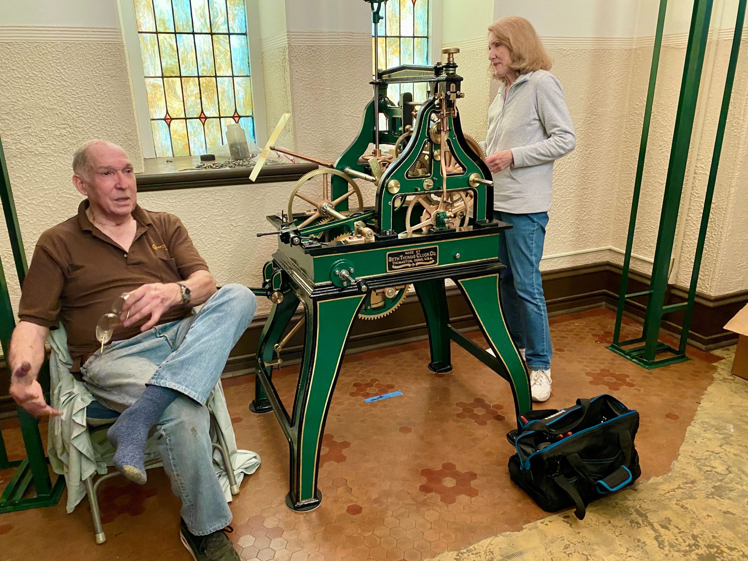 Man sitting in chair and woman standing at machine inside a lobby with stained glass windows behind them.