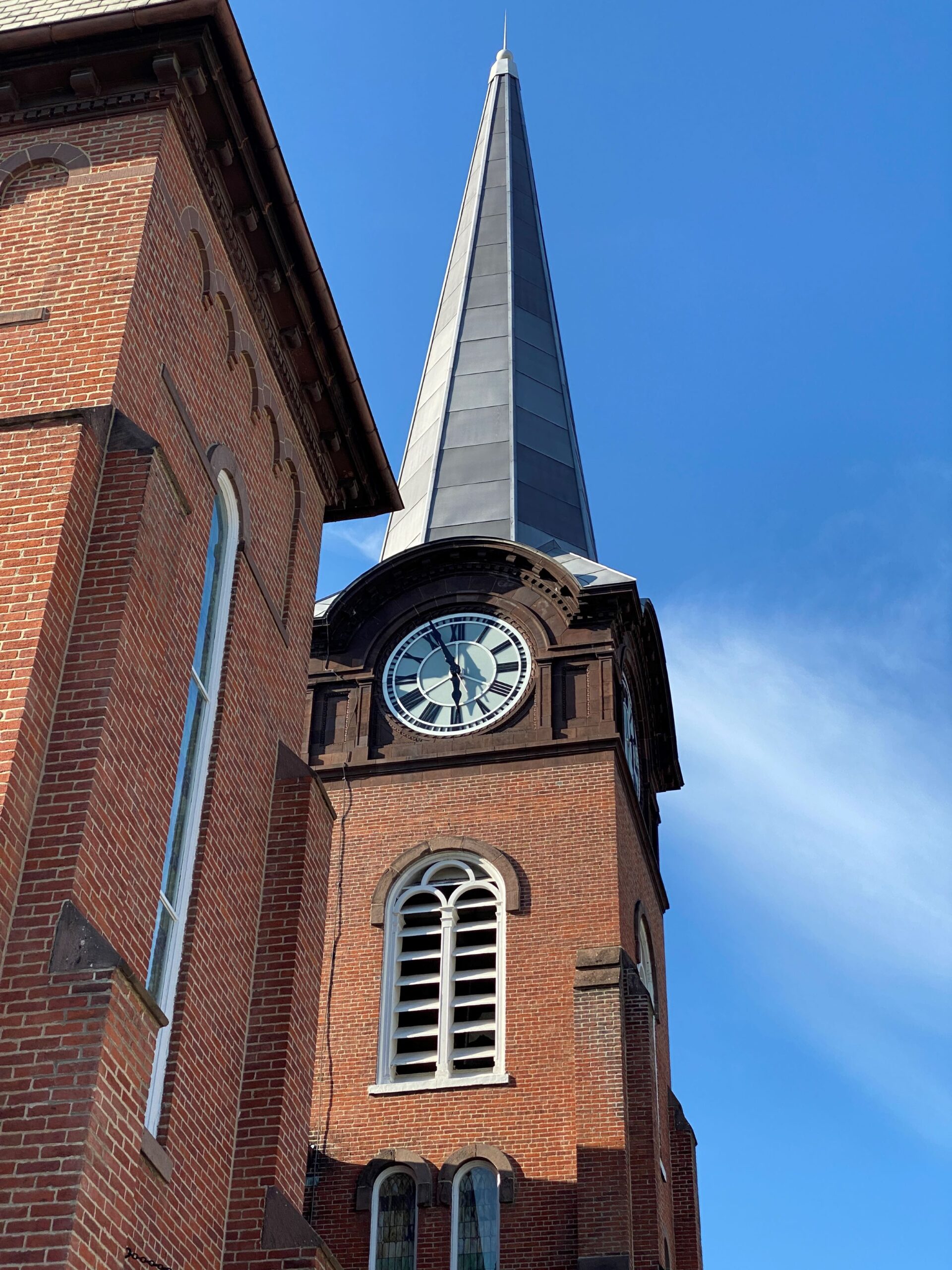 Looking up at a tall brick steeple with louvered window and clock face.
