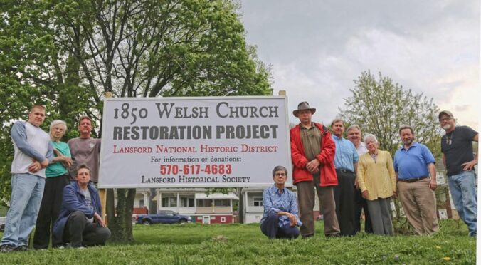 Group of men and women next to sign that says 1850 Welsh Church Restoration Project Lansford National Historic District.