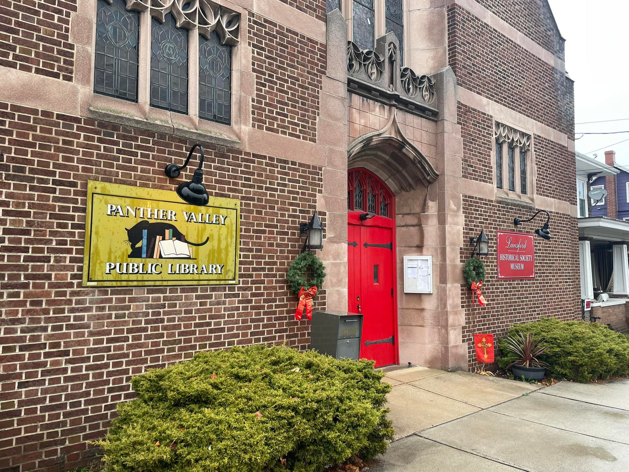 Red entrance doors in center of brick wall with stone details. Sign for Panther Valley Public Library left of door and sign for Lansford Historical Society Museum to right of door.