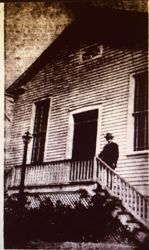 Man stands on steps in front of one story wood building with a center door and windows on either side.