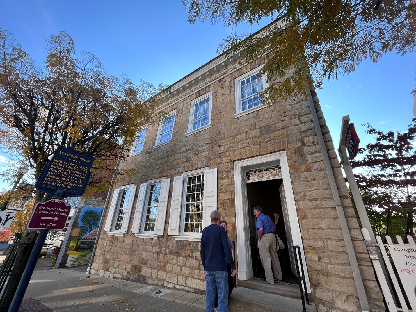 Three people stand at entrance to two-story stone house with many windows.