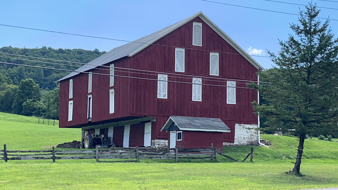 Large bank barn with many louvered windows on side and main sides surrounded by green grass and woods.