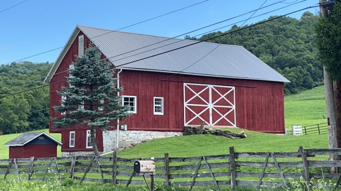Banked barn painted red and surrounded by green fields and woods.
