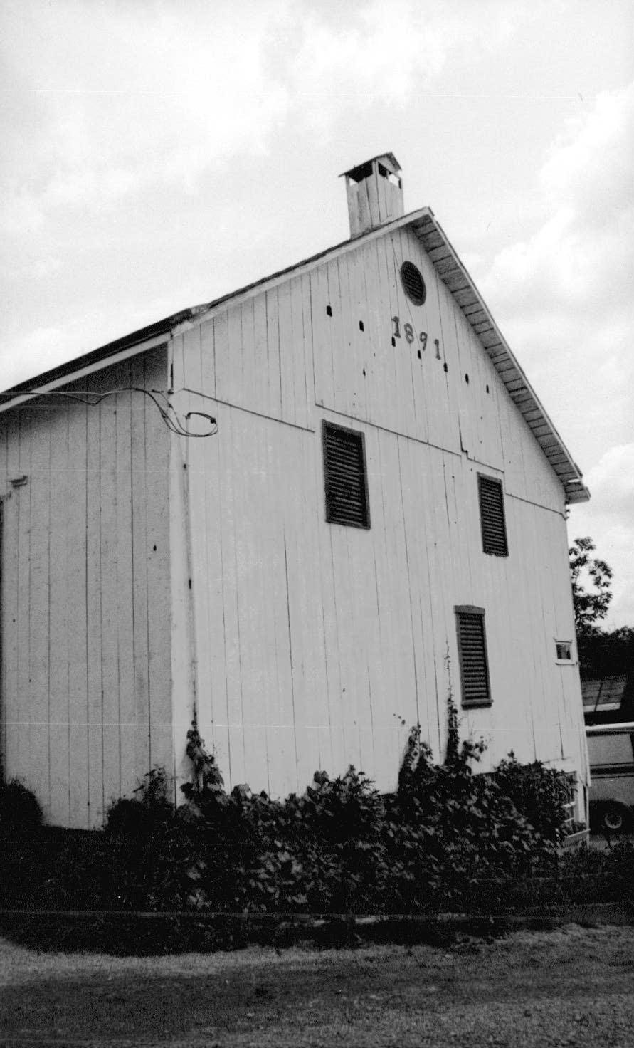 Two story building with a gable roof, vertical wood siding, louvered windows and 1891 in the gable.