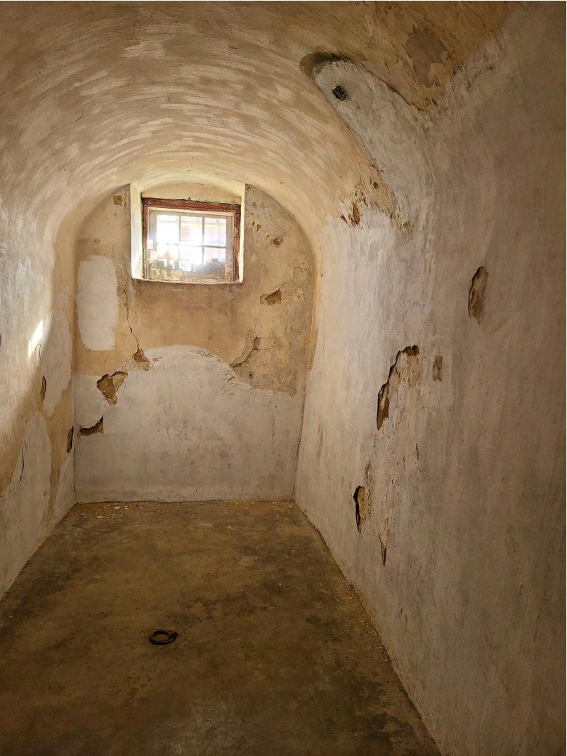 Small, narrow room with dirt floor, stucco/plaster-covered walls and arched ceiling, and a small window high on the back wall.