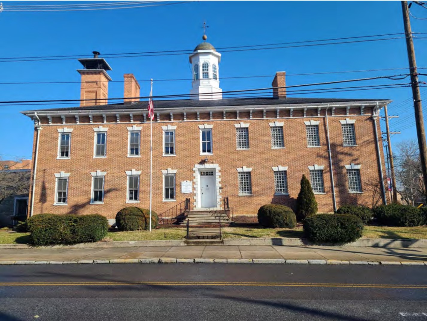 Large two-story brick building with center door with four window bays to each side and a roof with three chimneys and an enclosed cupola.