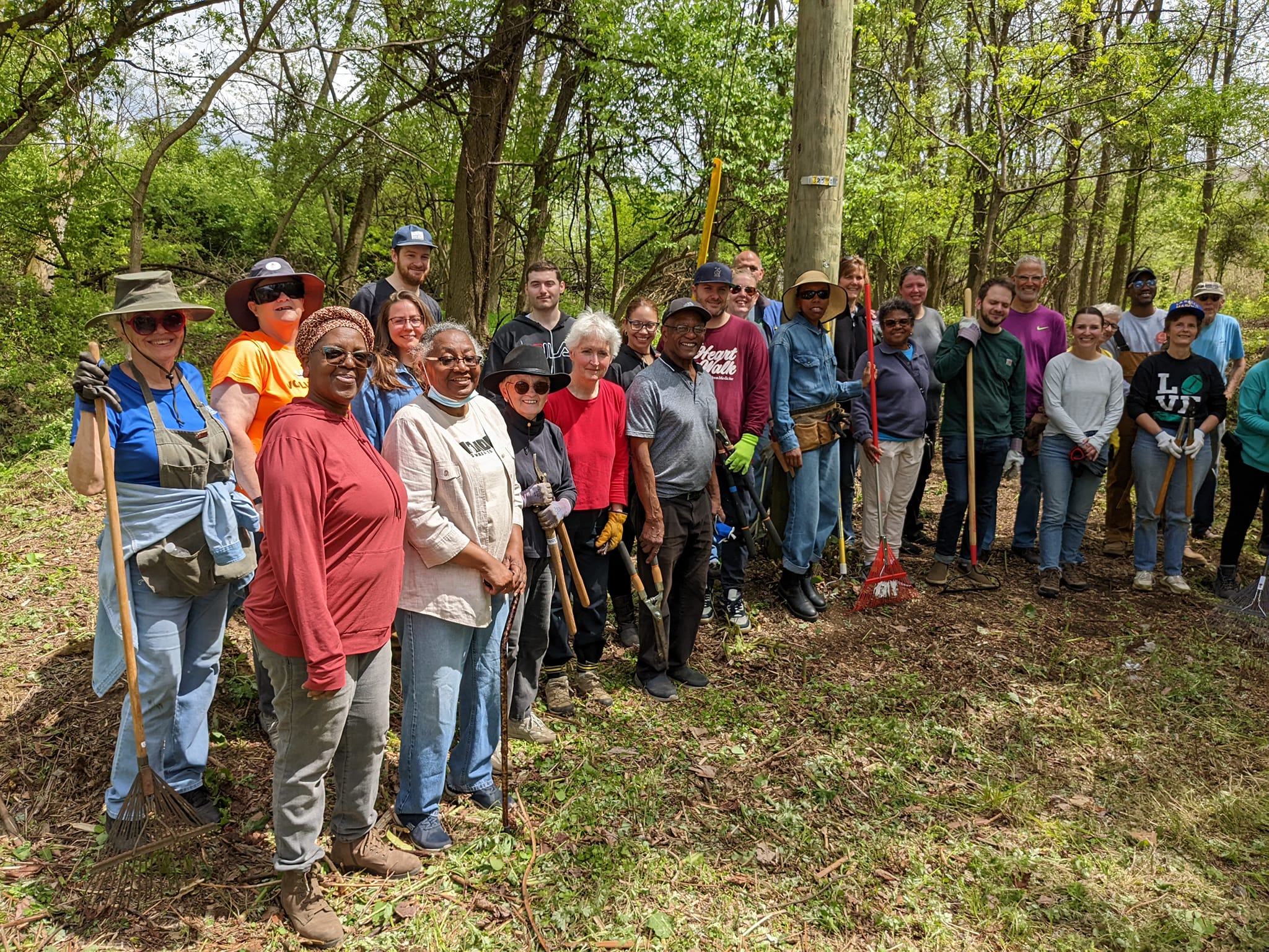 Large group of people standing outside in a wooded area and holding rakes and other landscaping tools.