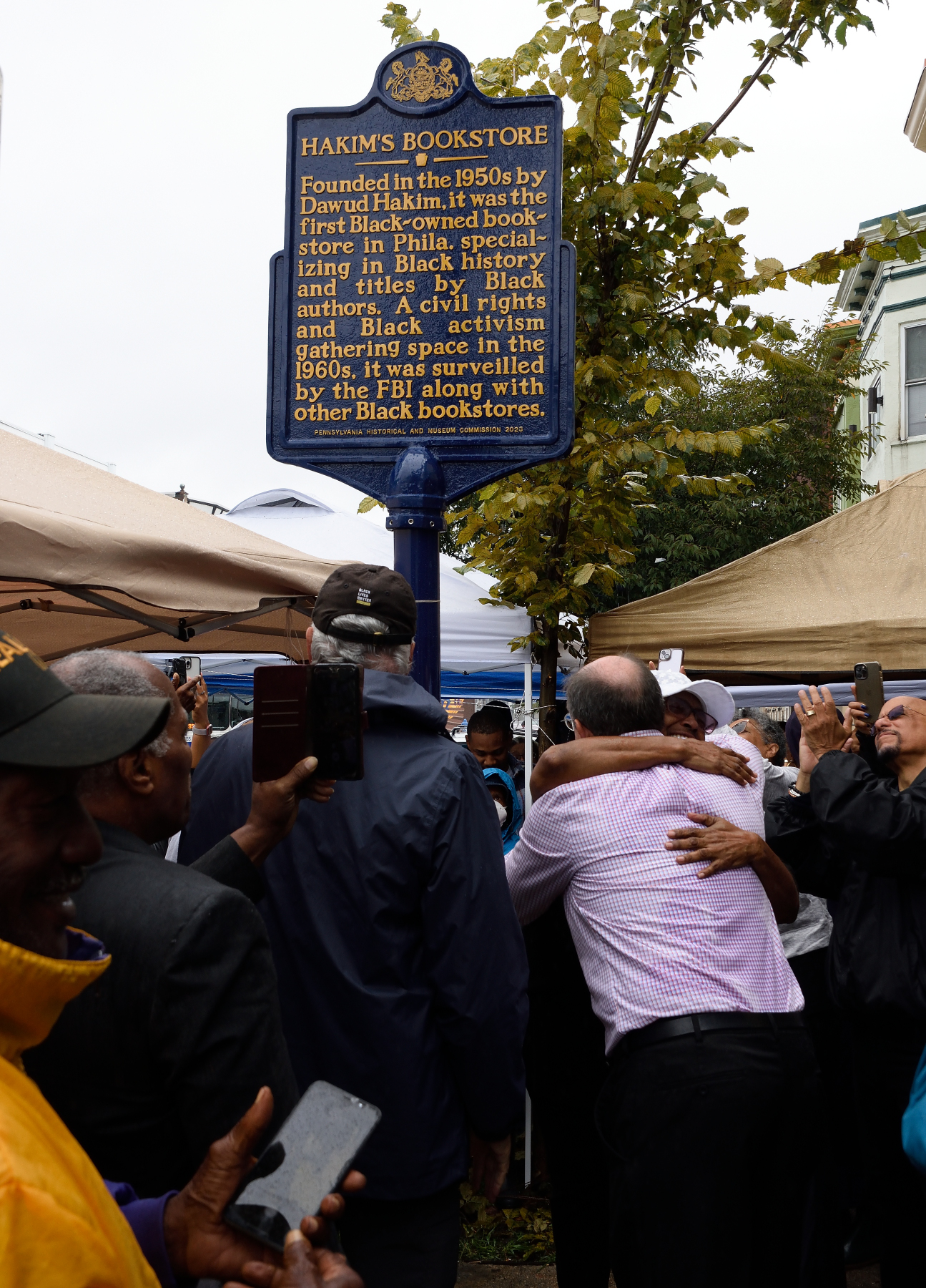 Large group of Black and white people stand outside around a tall metal pole with text panel on top.