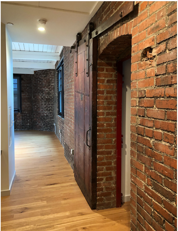 Narrow passageway with unpainted brick wall to right with an opening and to another space.