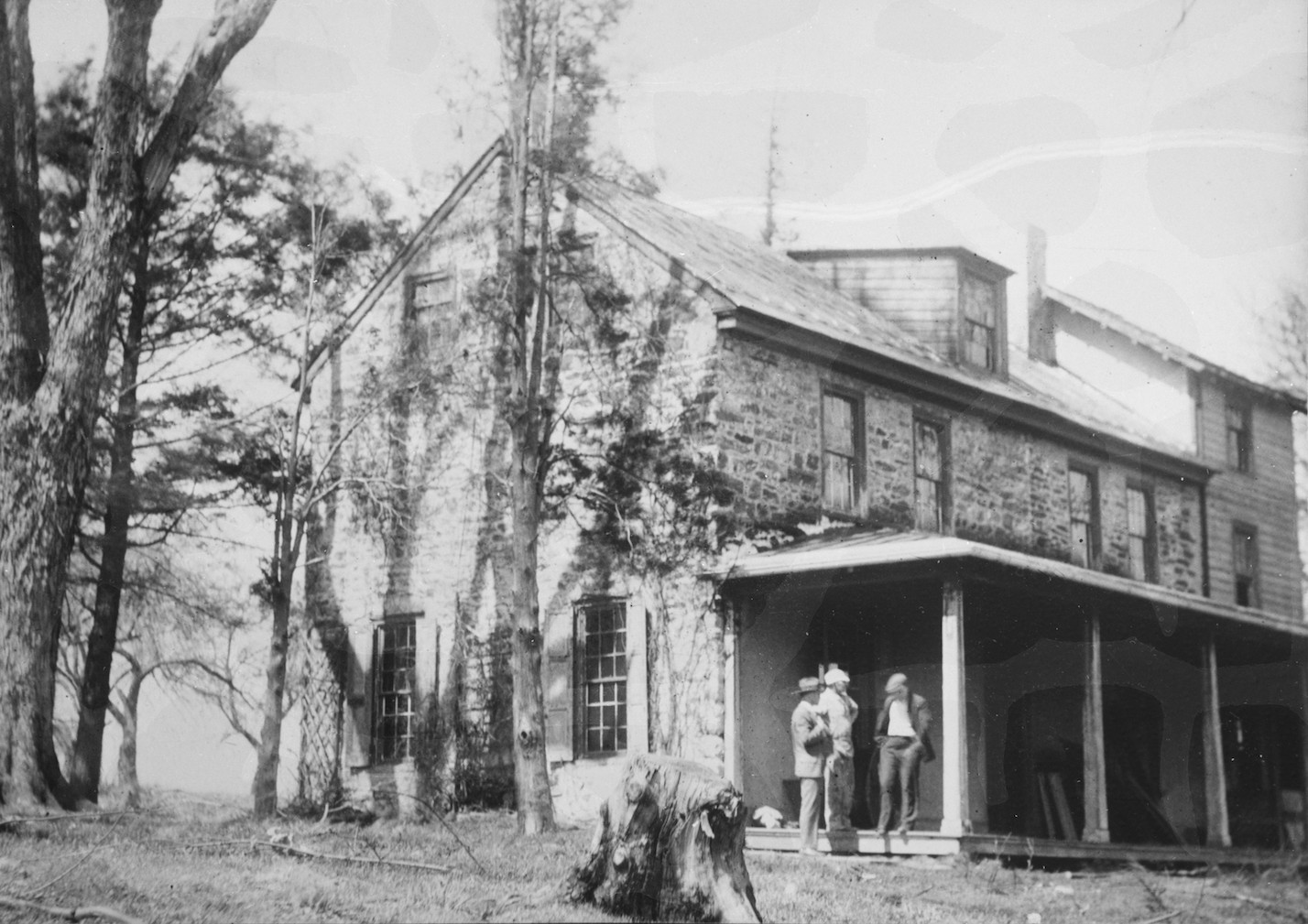 Black and white photograph of three men standing at the end of a wood porch attached to a two-story stone house.