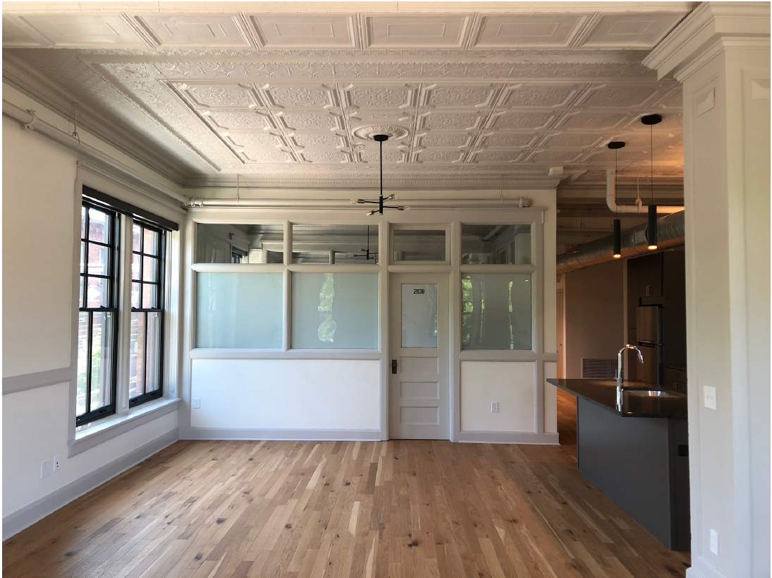 Room with wood floor, two windows in exterior wall at left, kitchen appliances to right, and wall with solid panels, door, and windows at back.