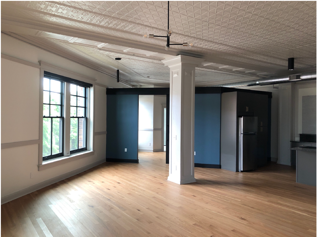 Large room with wood floor, two windows in exterior wall to left, decorative wood column, and kitchen appliances.