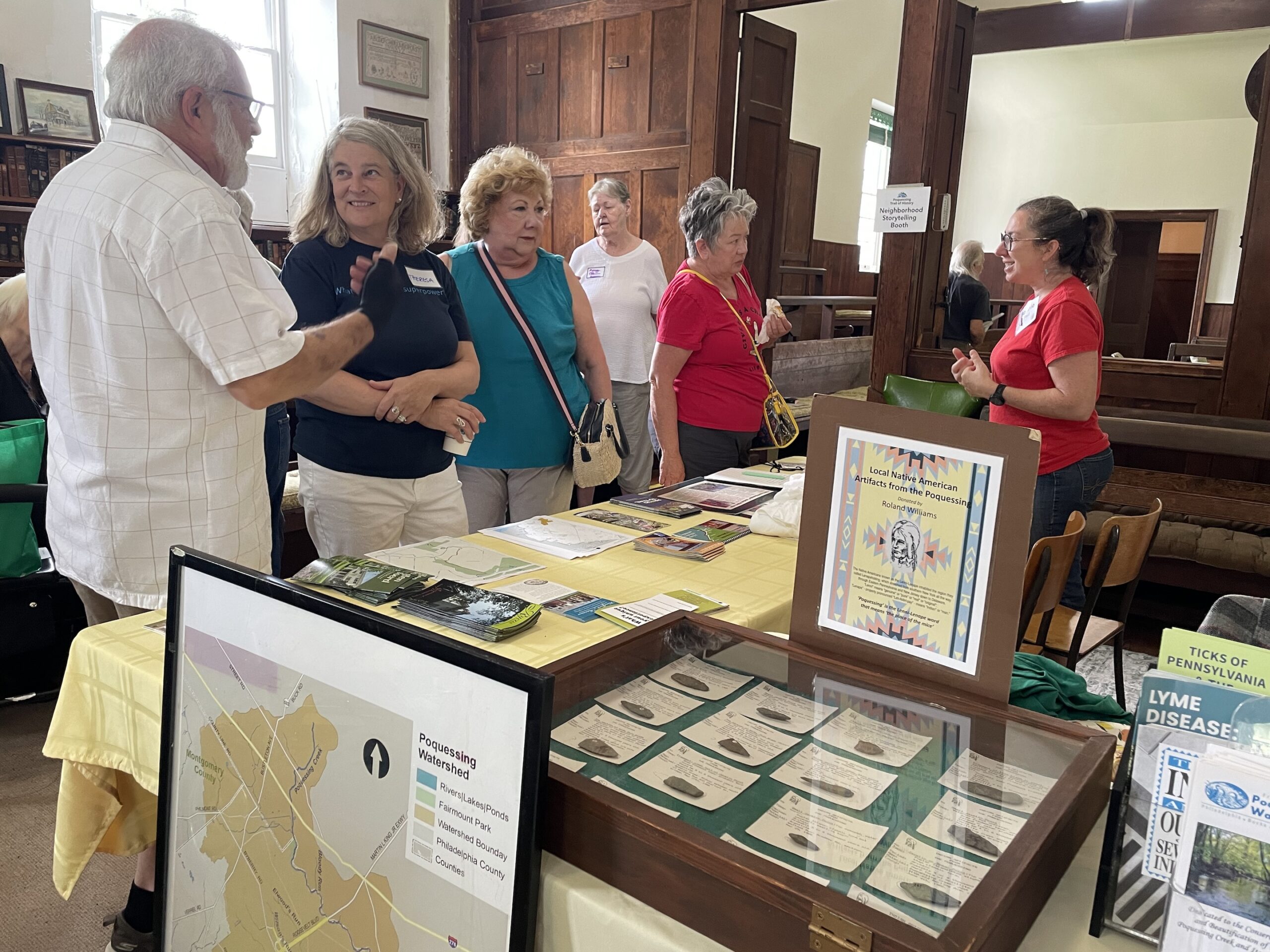 Group of five people stand talking at a table with printed papers and brochures inside a building.