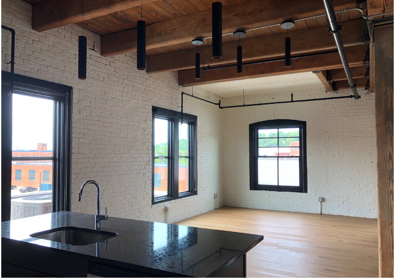 Room with windows in two exterior walls, painted brick, wood floor, wood ceiling, and small kitchen counter with sink.