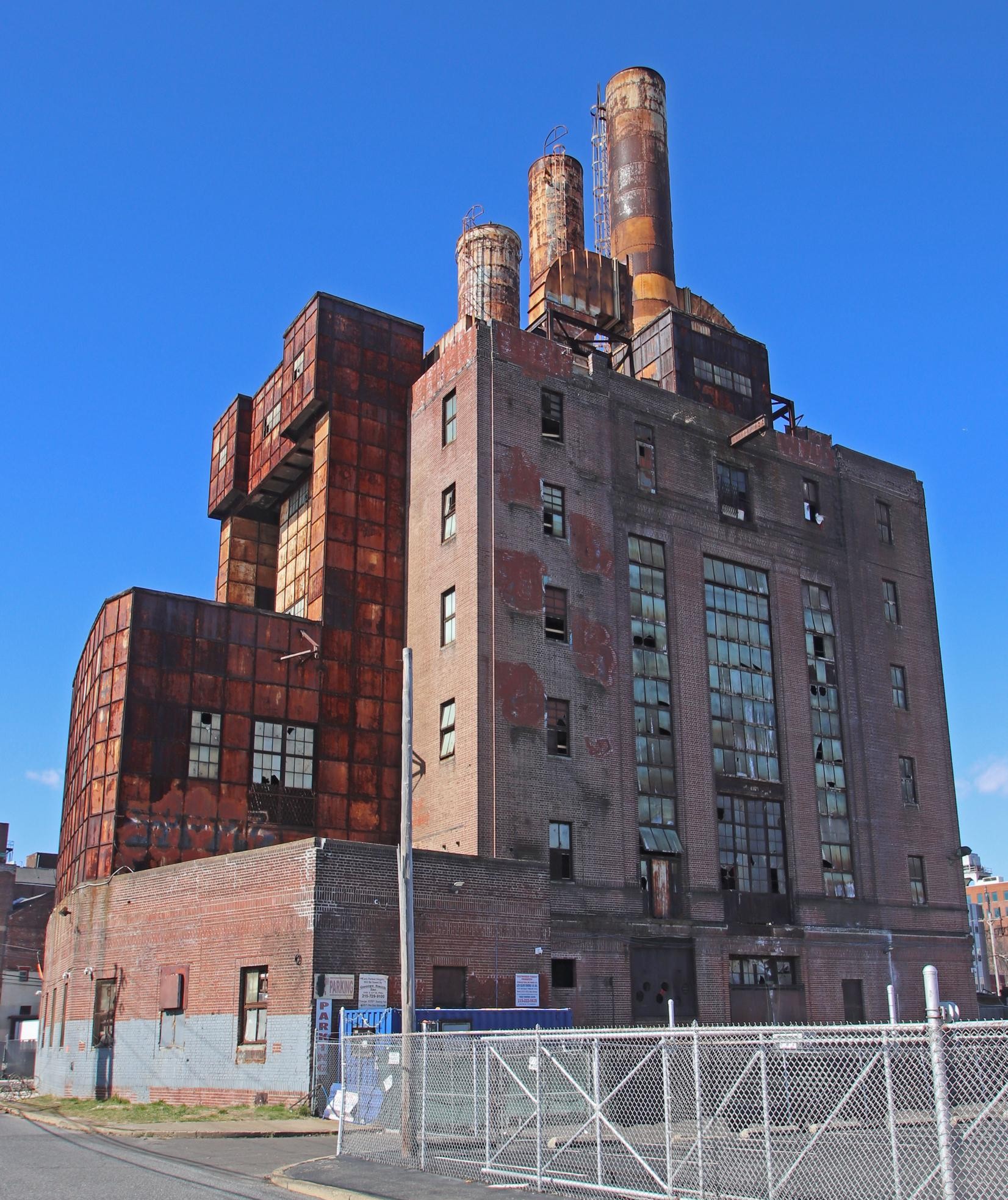 Very tall multi-story brick industrial building with three towers on roof and surrounded by pavement and fencing.