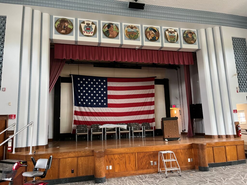 Shallow wood stage in a large room surrounded by curtains and plaster walls, with a large American flag on the stage.