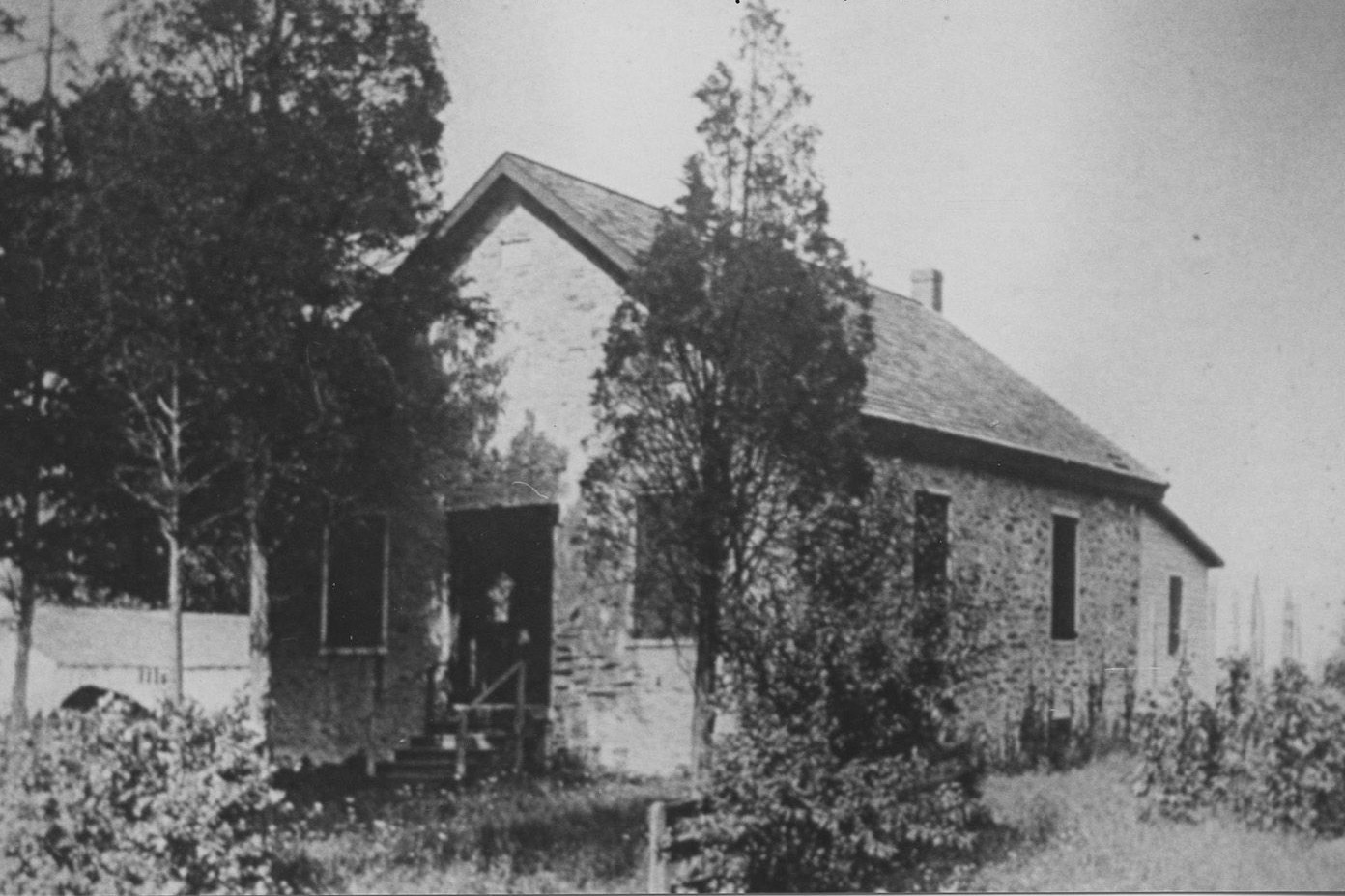 Black and white photograph of a one story stone building surrounded by trees and tall landscaping.