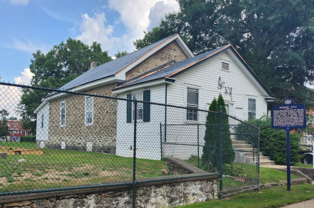 Small one story stone and wood building in a neighborhood setting with a blue metal marker in front and fenced yard to the side.
