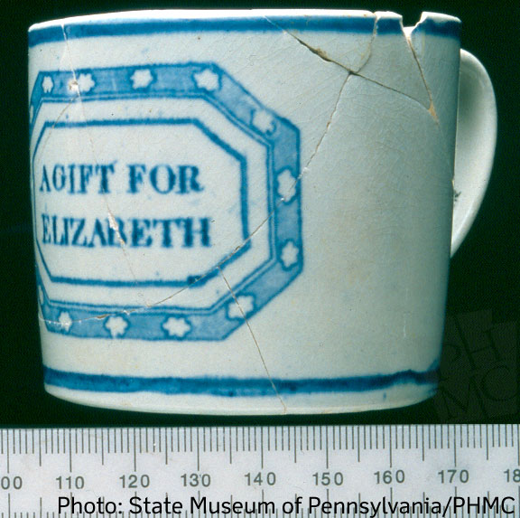 Small white ceramic cup with blue text and borders above a black and white ruler.
