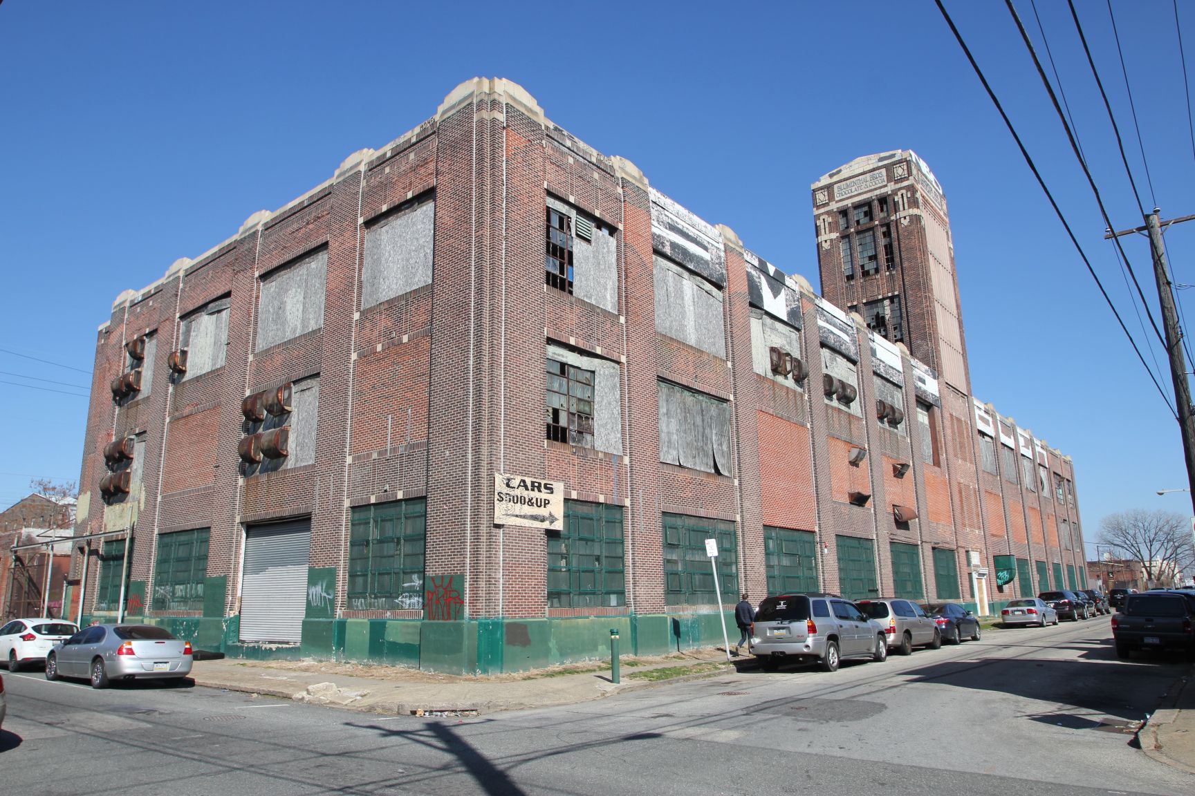Brick industrial building with very large broken metal and boarded up windows in an urban setting.
