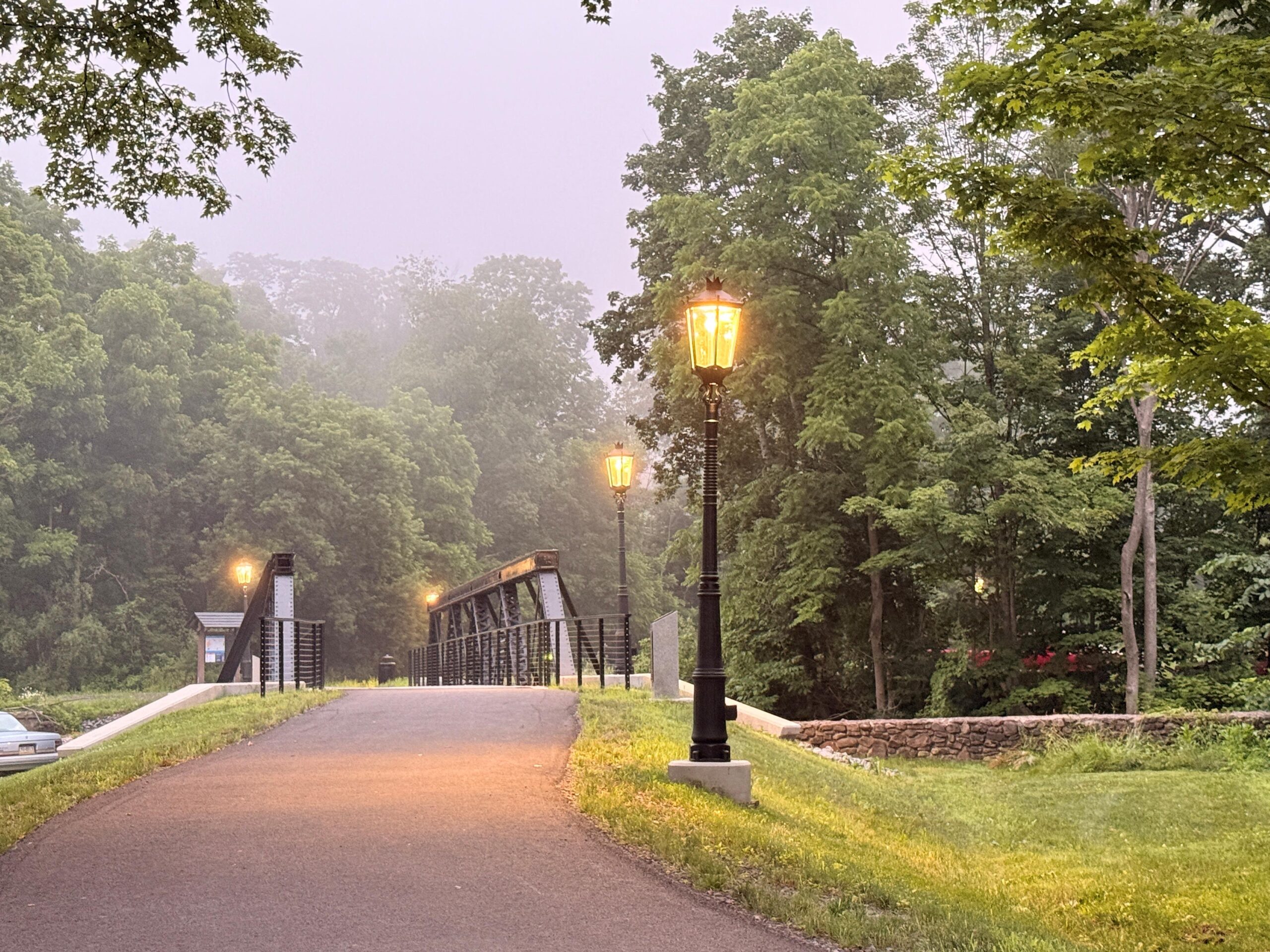 Paved path through grassy area leading to bridge with metal lampposts throughout.