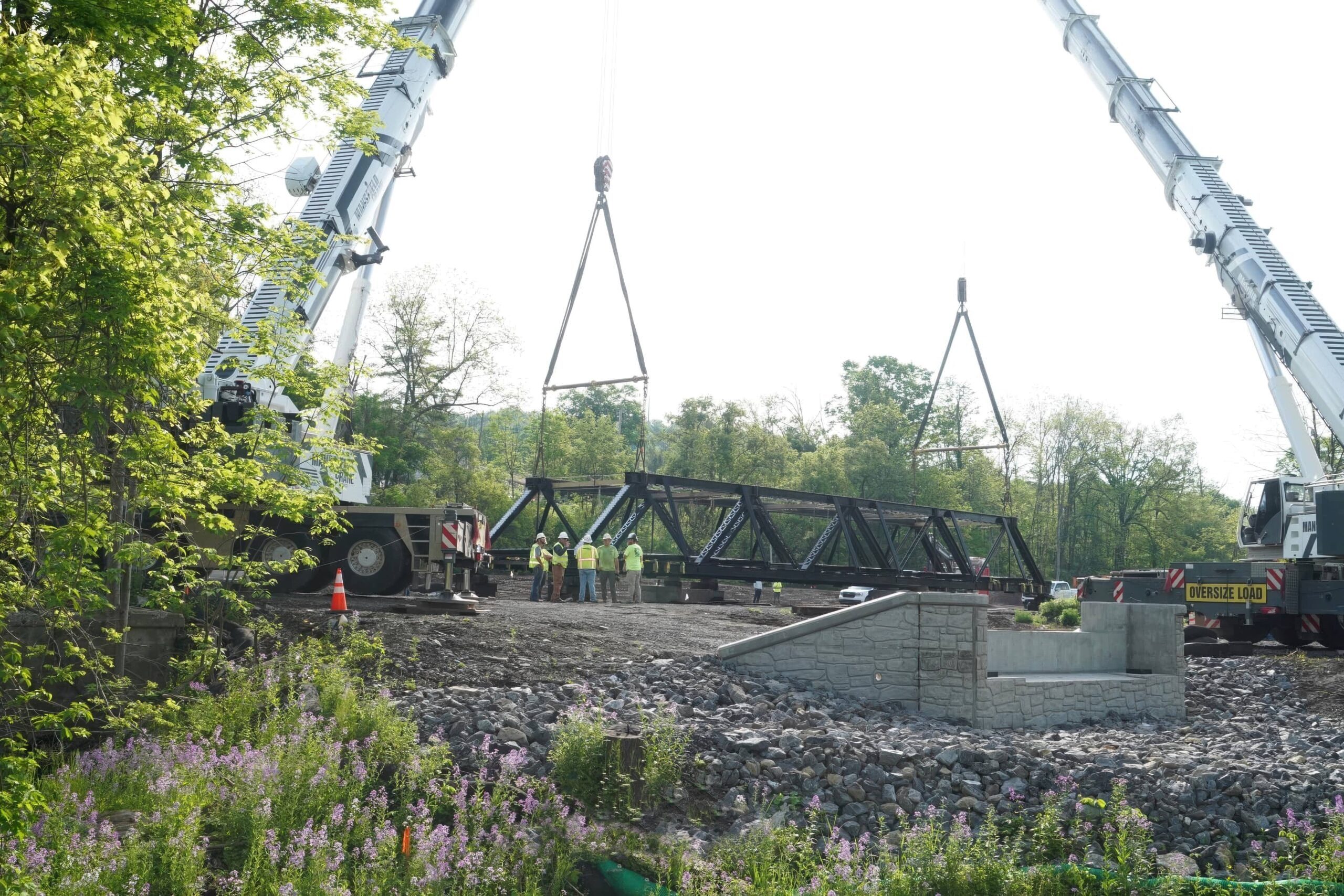 Two large cranes hold a large metal bridge on a worksite.