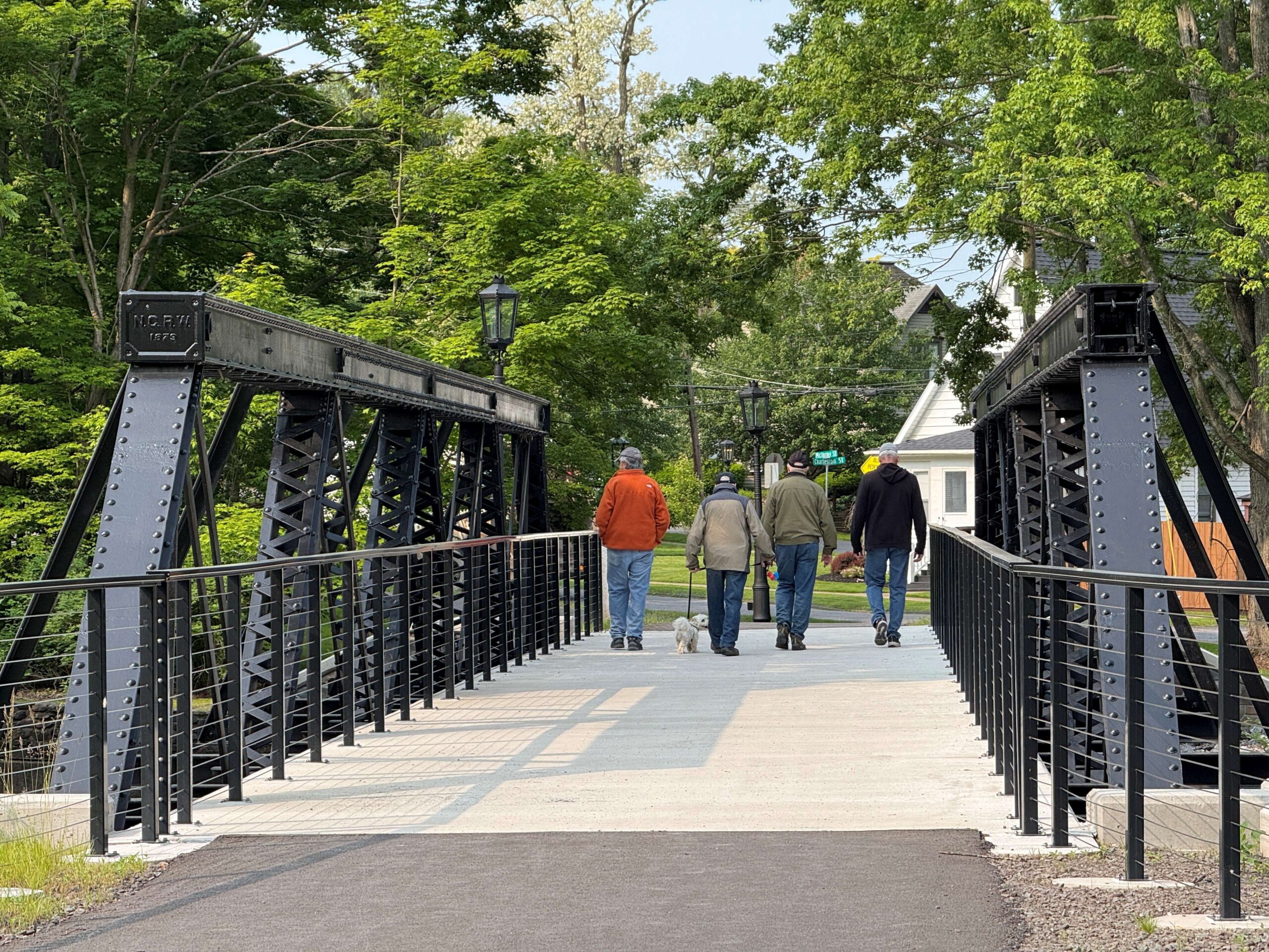 Group of four people and a dog walk across a paved path on a metal bridge.