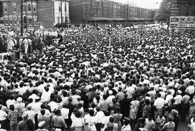 People crowded on a city street between rows of buildings.
