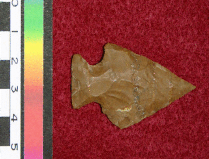 A Perkiomen style projectile point, made out of a reddish brown stone. The right side corner of the projectile head is broken and rounded off.